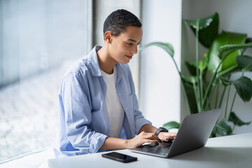 Fototapeta premium Focused latin millennial woman typing on laptop at white table, with smartphone and plants