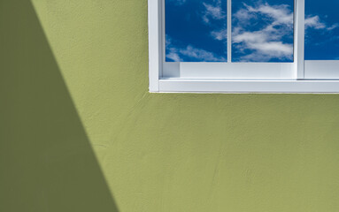 Green concrete wall with shadow light overlay and white window frame through blue sky