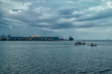 Fishings boat floats in the middle of the sea with a port and a large cargo ship in the background.