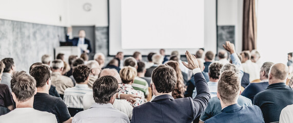 I have a question. Group of business people sitting at the chairs in conference hall. Businessman raising his arm. Conference and Presentation. Business and Entrepreneurship. - obrazy, fototapety, plakaty