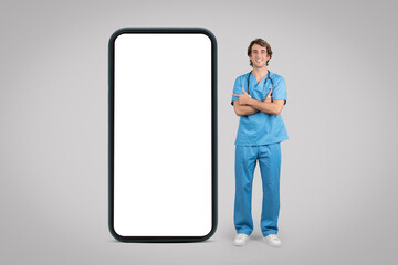 Cheerful man nurse, surgeon in blue uniform point finger at big phone with empty screen, isolated on gray background
