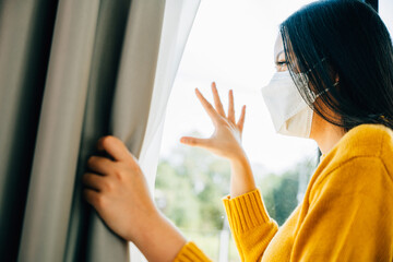 An upset Asian woman wearing a protective mask looks through the window feeling distressed and...