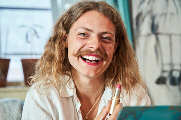 Portrait of the laughing longhaired guy with moustaches holding pink lipstick and looking at camera