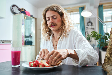 Longhaired man with makeup eating croissant with strawberry while having breakfast