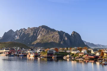 Foto auf Alu-Dibond Evening light bathes the scenic town of Reine in Lofoten, Norway, with its iconic rorbu cabins by the calm waters, framed by the imposing craggy peaks © Artem