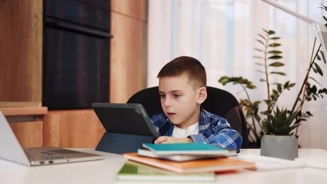 Little guy engaging in remote schooling through digital tablet while achieving useful skills and knowledge. Clever boy making research for his school project while sitting at home desk with books.