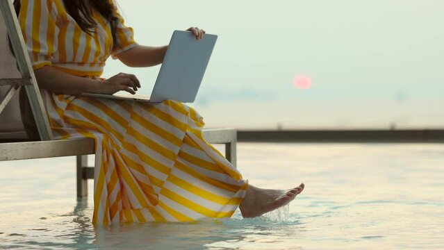 Closeup of Hands using and typing on laptop while sitting on a chair in the pool. Sea and bright sunset. A tropical beach for relaxation and travel.