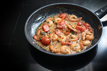 Shrimps in tomato cream sauce sauteed with herbs, onions and garlic in a black frying pan on the stove top, cooking a Mediterranean seafood meal, selected focus