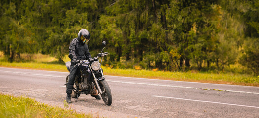 motorcyclist in motorcycle clothing and a helmet on a custom stylish motorcycle on a forest asphalt road - obrazy, fototapety, plakaty