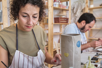 Focused female potter carefully applying colorful glaze to a decorated ceramic cylinder in an art...