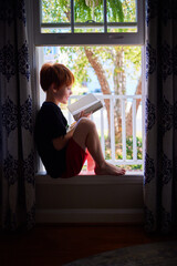 young boy enjoys reding a book while sitting in open window on sunny day