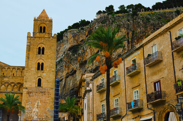 Landscape view of old city in Cefalu. Tower of ancient Cathedral of Cefalu and old residential house. Rocky mountain with green trees in the background. Travel and tourism concept. Cefalu, Sicily