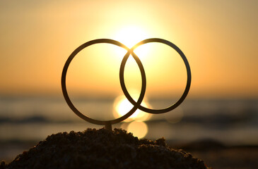 Black outlines of wedding rings at dawn and sunset on seashore. Stick contours in shape of wedding rings in sand on backdrop of setting and rising sun. Concept wedding love infatuation Valentine's Day - obrazy, fototapety, plakaty