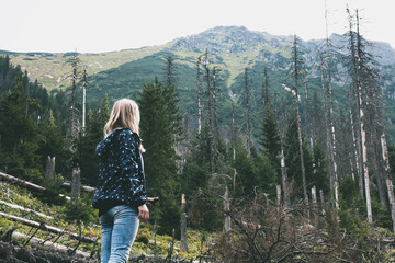 Young blonde girl looking at fallen trees in spring forest on background of mountains
