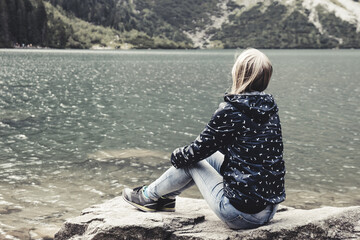 Young lonely blonde girl sitting in a romantic sadness on the shore of a blue lake in the mountains - obrazy, fototapety, plakaty
