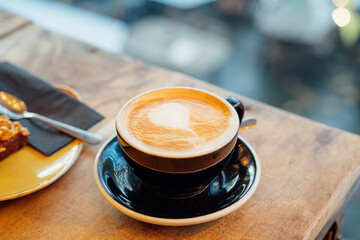 Close up hot cappuccino ceramic black coffee cup with heart shape latte art on the wooden craft table at cafe shop. Warm and cozy food and drink concept. Soft selective focus