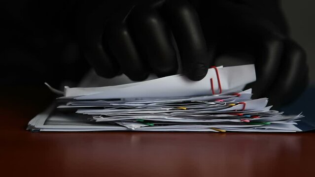 An unknown man in black gloves is rummaging through other people's documents lying on the table. A criminal is going through a folder of papers in someone else's office.