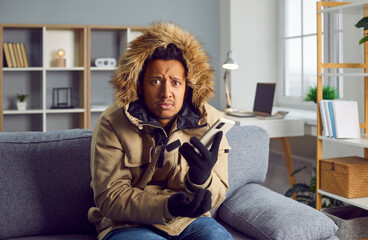 Fototapeta premium Young man is cold inside the house in winter. African American man wearing a warm parka jacket sitting in the living room with a very low temperature and making a phone call to the heating services