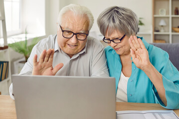 Happy senior family couple video calling relatives. Old man and woman in eyeglasses sitting at table with laptop computer, looking at screen, waving hands, saying hello and smiling