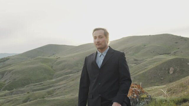 Portrait of an adult young man in a suit with a tired and haggard face against the background of a sunset in the mountains. The wind blows his hair. Unique landscape. Slow motion