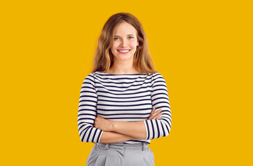 Studio portrait of happy beautiful woman in casual wear. Young blonde long haired girl in striped top standing with her arms crossed isolated on orange yellow background, looking at camera and smiling