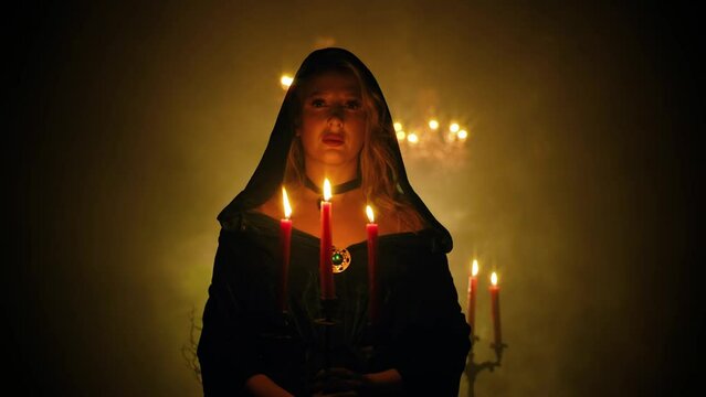 Medieval fantasy woman walking in dark room holding old candlestick in hands candles burning magical warm light. Vintage style cape dress with hood . Sexy girl vampire blows extinguishes candle fire