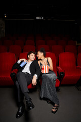 happy young diverse couple in evening attires holding popcorn and soda and smiling at each other