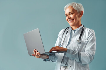 Beautiful friendly mature grey haired female doctor in white medical clothes holding a laptop and looking at the screen smiling while standing on a blue background. Doctor's appointment and services.