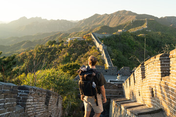 Joven viajero de espaldas en la muralla China al atardecer, en un paisaje montañoso - obrazy, fototapety, plakaty