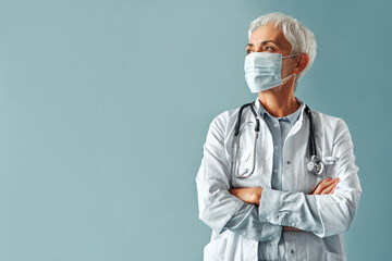 Mature confident short haired gray haired female medic in white coat, stethoscope around neck and mask looking sideways at free space blue background. Advertising of medical services, diagnostics.