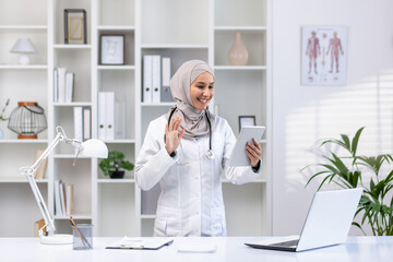 A smiling Muslim doctor in a hijab conducts an online appointment with a patient, holds a tablet,...