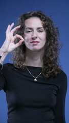 Woman giving OK sign with hand, approval gesture looking directly at camera standing on blue...