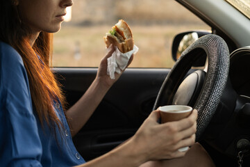 A happy woman driver is drinking coffee and eating a burger while driving a car. Concept of ordering fast food delivery