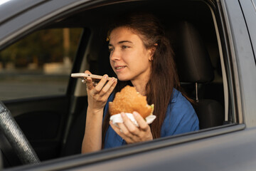 Focused woman sitting in car when recording voice message on smartphone. Busy lady sending voice message on cellphone and eating burger in car.