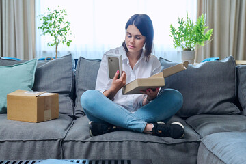 Young woman sitting at home unpacking cardboard box with online shopping - obrazy, fototapety, plakaty
