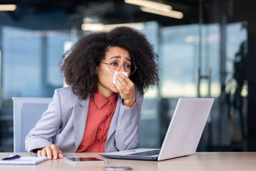 African American woman working in the office has a runny nose, blows her nose into a tissue, sits...
