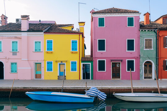 The vividly painted houses of Burano, Venice reflect in the still canal water, under a clear blue sky