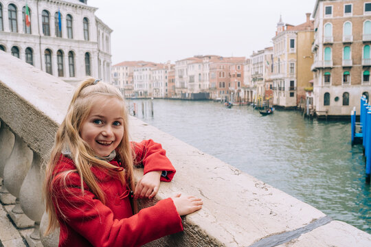 Young girl in a vivid red jacket rests her arms on a stone balustrade, overlooking the grandeur of a foggy Venetian canal