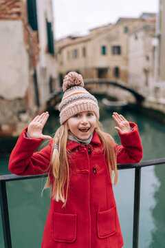A cheerful young girl in a red coat and striped beanie poses with an animated expression on a Venetian bridge, canal in the background