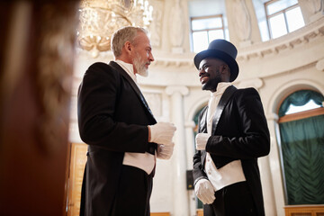 Low angle view at two gentlemen wearing classic tailcoats enjoying conversation in palace hall,...