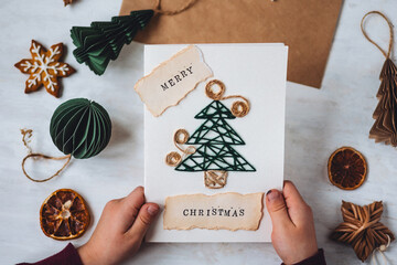 Concept of Christmas craft, handmade presents, family leisure time, hobby, recreation. Little kid with paper postcard with new year fir tree embroidering. Gingerbread cookies, lights, cozy atmosphere - obrazy, fototapety, plakaty