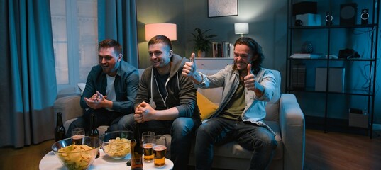 Male football fans sitting on couch in front of TV screen and cheering for favorite team on Euro2024 with beer and snacks at night. Guys celebrating win in match and hugs while spending time together.