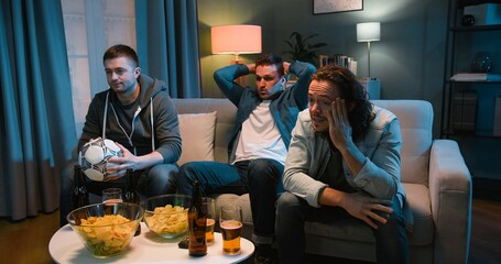 Handsome Caucasian men football fans sitting on couch and watching football match late at night....