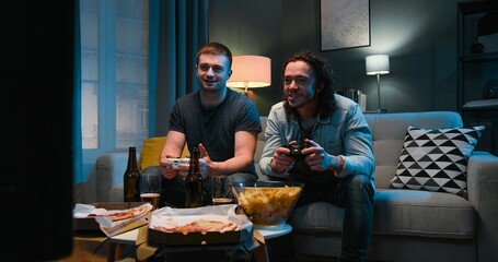 Two Caucasian friendly guys playing videogame with joystick while sitting in tension late in evening with snacks and beer in front of TV. Men having fun together with games.