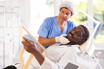 Experienced male cosmetologist examines facial skin of African-American man patient and advises on...