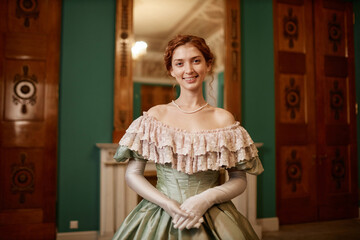 Waist up portrait of elegant young lady wearing ballgown posing in palace hall and looking at...