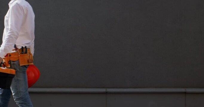 Builder walks across site ready for task ahead with helmet and tool-belt securing well-equipped toolbox. Readiness of service employee for task