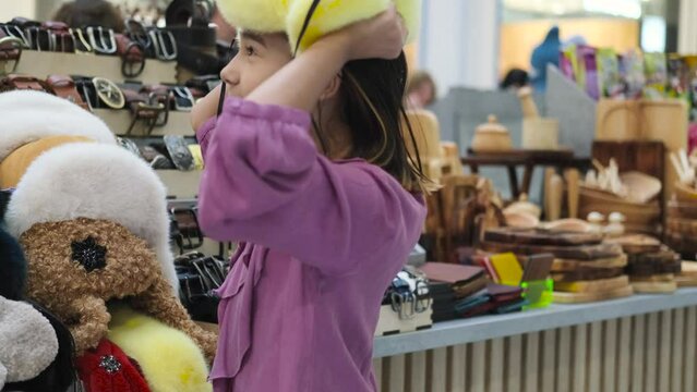 A little girl tries on a bright yellow fur hat with earflaps at a fair. Clothes for cold winter weather. Warm clothes and fashionable accessories for children.