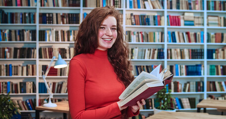 Close-up portrait of red head geek girl preparing for final test and reading book in library. Bookcase in background.