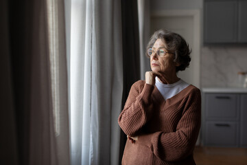 Retirement and depression. Upset thoughtful senior woman standing and looking at window at home,...